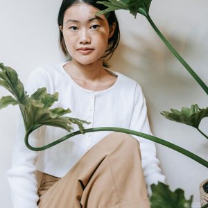 Woman in white cardigan sitting on white floor with plant.